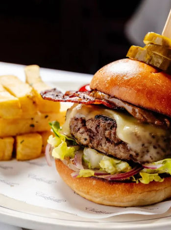 Gourmet cheeseburger on a brioche bun with melted cheese, juicy beef patty, crispy bacon and pickles, lettuce and red onion, served with thick-cut seasoned fries on a white plate