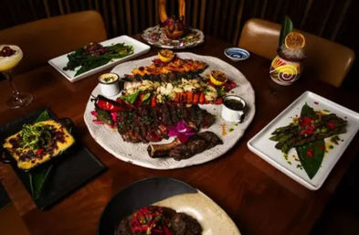 Restaurant table spread featuring a large surf-and-turf platter with grilled steak, lobster tails, shrimp skewers, colorful garnishes, roasted vegetables, asparagus sides and cocktails