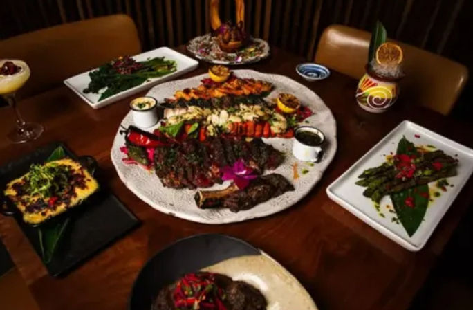 Restaurant table spread featuring a large surf-and-turf platter with grilled steak, lobster tails, shrimp skewers, colorful garnishes, roasted vegetables, asparagus sides and cocktails