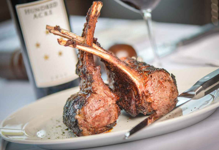Charred herb-crusted lamb chops stacked on a white plate with a steak knife and a blurred wine bottle and glass in the background, upscale restaurant entrée.