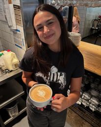 Smiling barista holding a large latte with heart latte art at a cozy cafe counter, espresso equipment and stacked saucers in the background