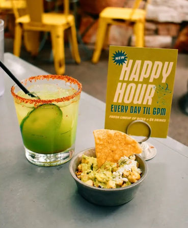 Chili-rimmed cucumber margarita next to a bowl of guacamole with a tortilla chip and a 'Happy Hour every day til 6pm' sign on a bar table