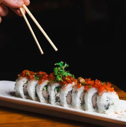 Hand holding chopsticks above a neatly sliced sushi roll topped with diced red pepper and microgreens on a white rectangular plate over a wooden table against a black background