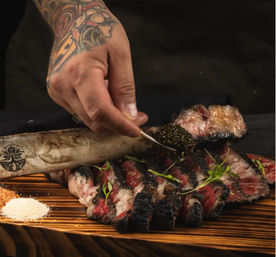 Tattooed hand spooning caviar onto bone‑marrow-topped charred sliced steak with microgreens and coarse salts on a rustic wooden board, gourmet plating