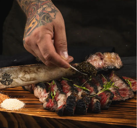 Tattooed hand spooning caviar onto bone‑marrow-topped charred sliced steak with microgreens and coarse salts on a rustic wooden board, gourmet plating
