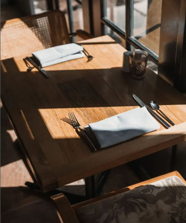 Sunlit wooden cafe table for two by a window, neatly set with folded napkins, forks, knives and spoons, wicker chairs and warm cozy brunch vibe.