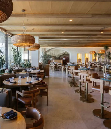 Spacious, airy restaurant interior with exposed wooden beam ceiling, woven pendant lights, round wooden tables set with blue napkins and glassware, leather chairs and bar stools, potted plants and large floor-to-ceiling windows bathing the space in warm natural light.