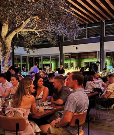 Lively covered patio restaurant at night with a large illuminated tree overhead, groups of friends dining at communal wooden tables, cocktails on the table and a glowing bar in the background, vibrant urban nightlife scene.