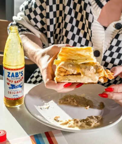 Flaky biscuit sandwich stuffed with fried chicken and creamy white gravy, held over a plate next to a bottle of hot sauce; person in a black-and-white checkered sweater with bright red nails.