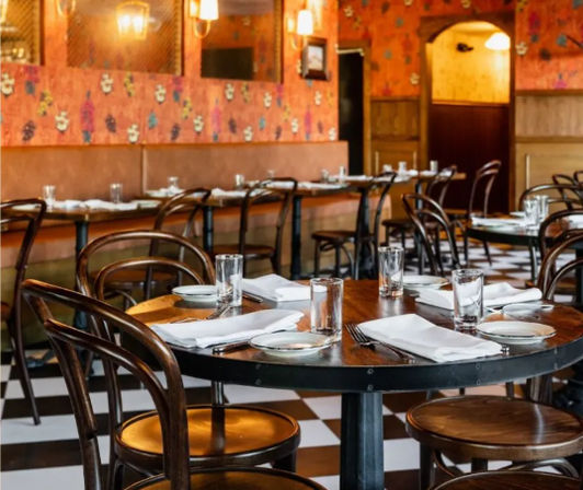 Cozy vintage bistro interior with round wooden tables and bentwood chairs, black-and-white checkered floor, floral wallpaper, and tables set with glasses, plates and white napkins, ready for diners.