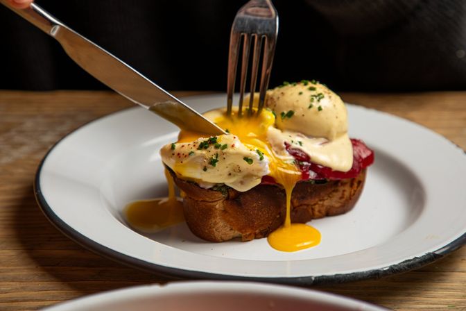 Close-up of eggs Benedict on toasted bread with poached eggs and hollandaise, fork and knife cutting a runny yolk spilling onto a white enamel plate on a wooden table — brunch close-up.