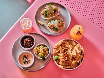 Top-down shot of a bright pink table with two fish tacos on a gray plate, three small bowls of salsa, guacamole and crema, a bowl of tortilla chips and fried bites, and two citrus cocktails.