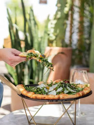 Hand lifting a slice of artisan arugula pizza with shaved Parmesan from a raised black pizza pan on a gold stand, outdoor patio table with potted cacti and a wine glass in the blurred background.