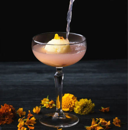 Pink sorbet cocktail in a coupe glass being poured with clear liquid, styled on a dark table with scattered yellow marigold flowers