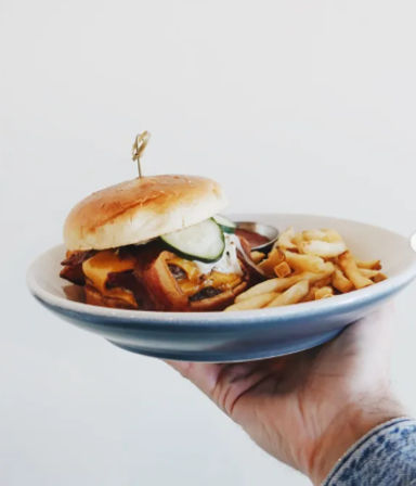 Close-up of a mouthwatering American bacon cheeseburger with melted cheddar and pickles, served with golden fries on a blue plate held by a hand against a white background.