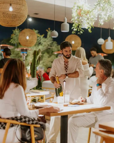 Server pouring champagne for diners at a lively outdoor patio restaurant at night, with warm rattan pendant lights, hanging greenery, and guests in white attire.