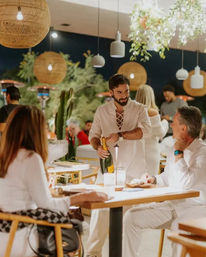Server pouring champagne for diners at a lively outdoor patio restaurant at night, with warm rattan pendant lights, hanging greenery, and guests in white attire.