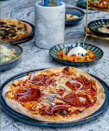 Artisan pepperoni pizza with melted mozzarella and red onion on a blue ceramic plate, set on a marble outdoor table with bowls of burrata and side dishes in the background.