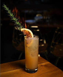 Tall citrus cocktail in a highball glass on a wooden table, garnished with a flaming rosemary sprig and charred citrus slice against a dim bar background.