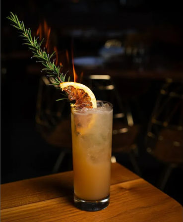 Tall citrus cocktail in a highball glass on a wooden table, garnished with a flaming rosemary sprig and charred citrus slice against a dim bar background.