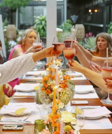 Friends clinking pink cocktails over a long brunch table with orange floral centerpieces, candles and place settings on a bright outdoor patio