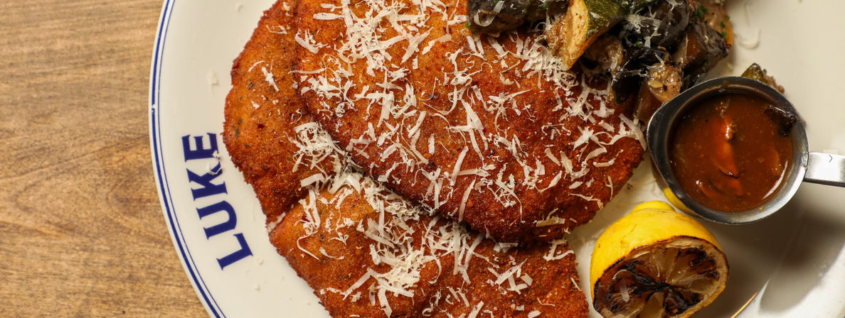 Golden-brown breaded cutlets topped with shredded Parmesan, paired with charred lemon, sautéed mushrooms and a small cup of savory sauce on a white plate over a wooden tabletop.