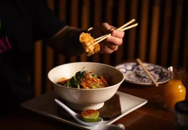 Hand using chopsticks to lift a dumpling from an Asian noodle bowl with greens and shredded carrots, served with lime, spoon, patterned side plate and a small bottle of orange juice on a wooden table.