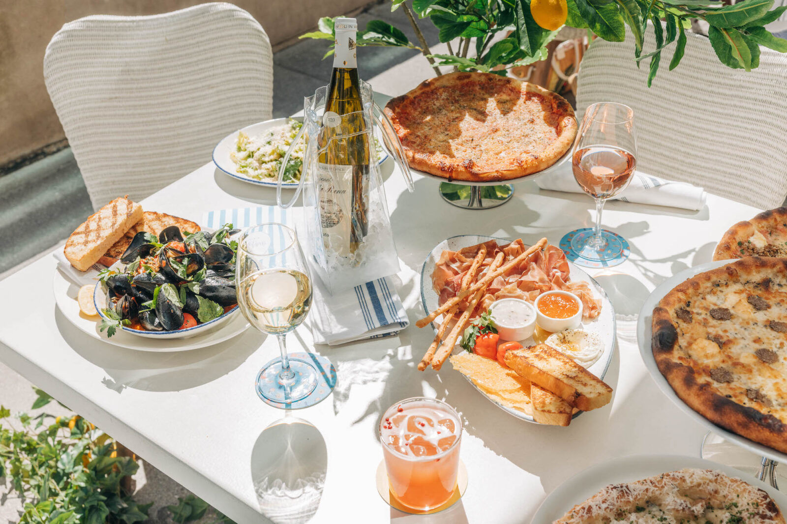 Sunlit al fresco table with wood-fired pizzas, bowl of mussels and grilled bread, charcuterie platter with breadsticks and dips, chilled white wine in an ice bucket, rosé and a pink cocktail