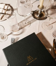 Fine-dining restaurant table with white tablecloth, crystal wine and water glasses, silverware, a lit candlestick, and a black leather menu embossed with a gold emblem.