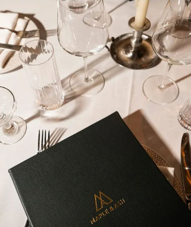 Fine-dining restaurant table with white tablecloth, crystal wine and water glasses, silverware, a lit candlestick, and a black leather menu embossed with a gold emblem.