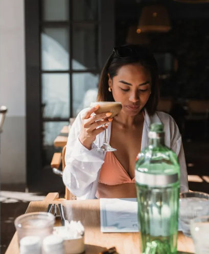 Woman in a coral bikini top and white shirt sipping a cocktail at a sunlit café table with a green bottle and menu — relaxed brunch vibe.