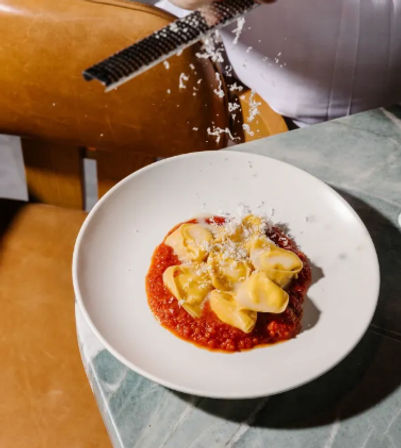 Parmesan flakes being grated over a plate of fresh tortellini on rich tomato sauce, served on a marble table.
