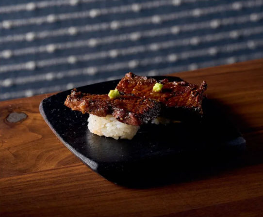 Charred grilled beef nigiri sushi topped with tiny wasabi dots on a black slate plate, set on a rustic wooden table with a blurred patterned background