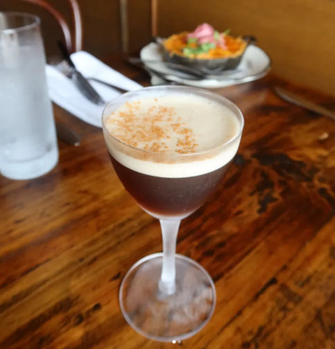 Espresso martini cocktail in a coupe glass with creamy foam and cocoa dusting, sitting on a rustic wooden table in a cozy restaurant with water glass and appetizer blurred in the background.