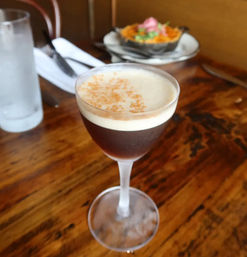 Espresso martini cocktail in a coupe glass with creamy foam and cocoa dusting, sitting on a rustic wooden table in a cozy restaurant with water glass and appetizer blurred in the background.