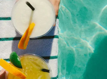 Overhead poolside shot of two tropical cocktails on a striped towel beside turquoise pool water — a frozen white drink and a citrus yellow cocktail garnished with orange peel and lime.