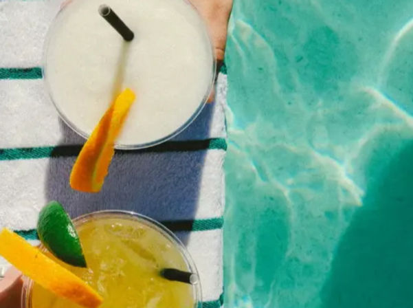 Overhead poolside shot of two tropical cocktails on a striped towel beside turquoise pool water — a frozen white drink and a citrus yellow cocktail garnished with orange peel and lime.