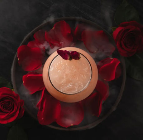 Top-down view of a pink frosty cocktail topped with dried rosebuds, surrounded by red rose petals and wisps of smoke on a dark tabletop — romantic floral drink.