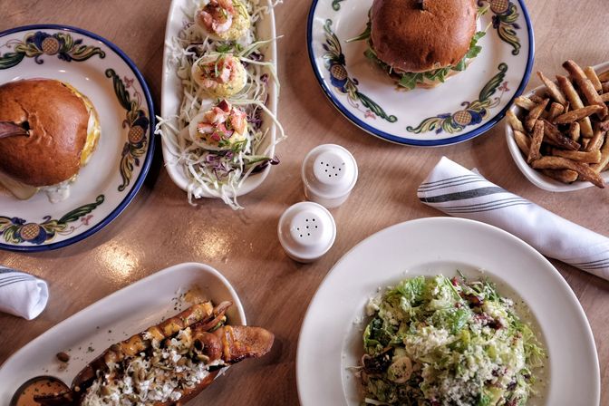 Overhead bistro-style brunch spread on a wooden table: two gourmet burgers on decorative plates, deviled eggs on shredded cabbage, a crisp green salad, a loaded sandwich with drizzle and seeds, a bowl of fries, and salt-and-pepper shakers.
