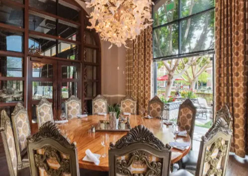 Elegant private dining room with a large round polished wood table surrounded by ornate upholstered chairs, a sculptural white chandelier overhead and floor-to-ceiling windows revealing a tree-lined outdoor patio