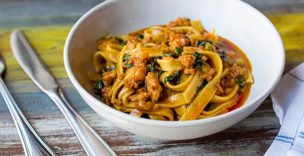 Hearty Italian tagliatelle with savory meat ragu and wilted greens in a white bowl on a rustic wooden table, with knife and fork beside it