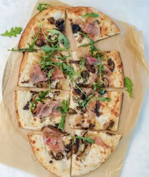 Overhead view of a rectangular artisan flatbread pizza on parchment, sliced into squares and topped with prosciutto, sautéed mushrooms, melted white cheese and fresh arugula