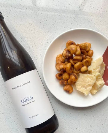 Craft pale ale bottle beside a white plate of toasted corn nuts and chunks of aged cheese on a speckled kitchen countertop.