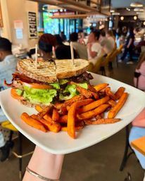 Grilled BLT on toasted sourdough with crispy bacon, lettuce and tomato, served with golden sweet potato fries on a white plate in a bustling café.
