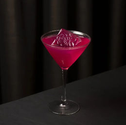 Vibrant magenta cocktail in a martini glass topped with a decorative rose-shaped ice cube on a black table with a dark curtain backdrop