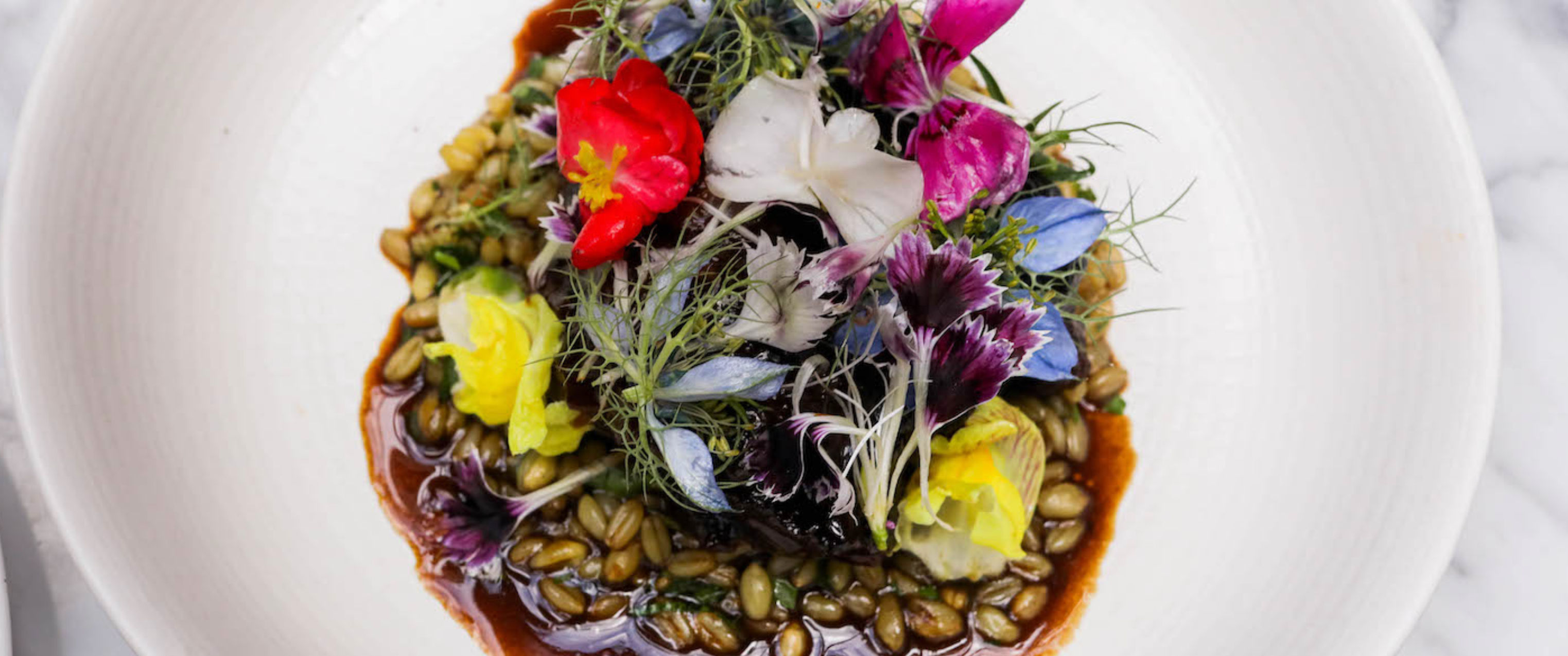 Top-down shot of a colorful gourmet grain bowl: toasted grains in a glossy savory sauce, topped with microgreens and vibrant edible flowers in a white ceramic bowl on a marble surface.