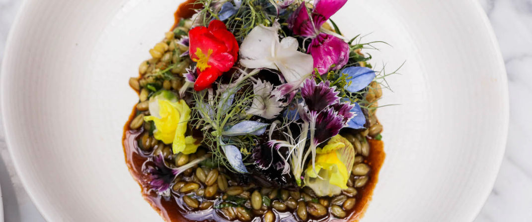 Top-down shot of a colorful gourmet grain bowl: toasted grains in a glossy savory sauce, topped with microgreens and vibrant edible flowers in a white ceramic bowl on a marble surface.