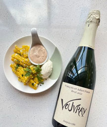 Plate of seasoned corn-on-the-cob pieces with creamy dipping sauce and herbs beside a chilled bottle of sparkling white wine on a light kitchen counter
