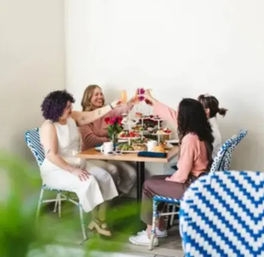 Four friends cheerfully toasting over brunch at a bright minimalist cafe table with a tiered tray of pastries, coffee, and blue bistro chairs