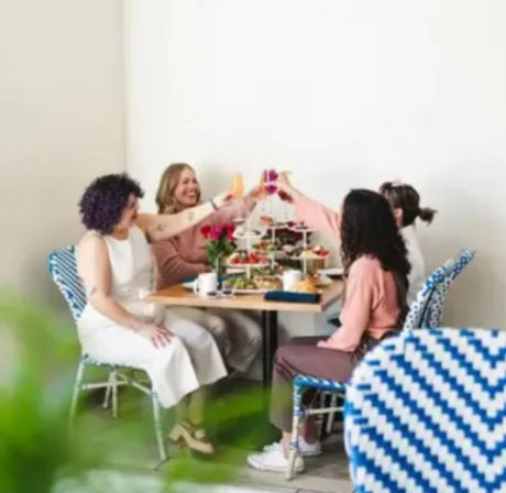 Four friends cheerfully toasting over brunch at a bright minimalist cafe table with a tiered tray of pastries, coffee, and blue bistro chairs